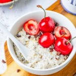 A white bowl filled with oatmeal, low carb chocolate, and a few cherries on a wooden cutting board.