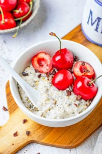 A white bowl filled with oatmeal, low carb chocolate, and a few cherries on a wooden cutting board.