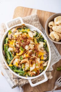 A white casserole dish filled with green beans and mushrooms that have been topped with melted cheese, crispy onions and pork rinds.