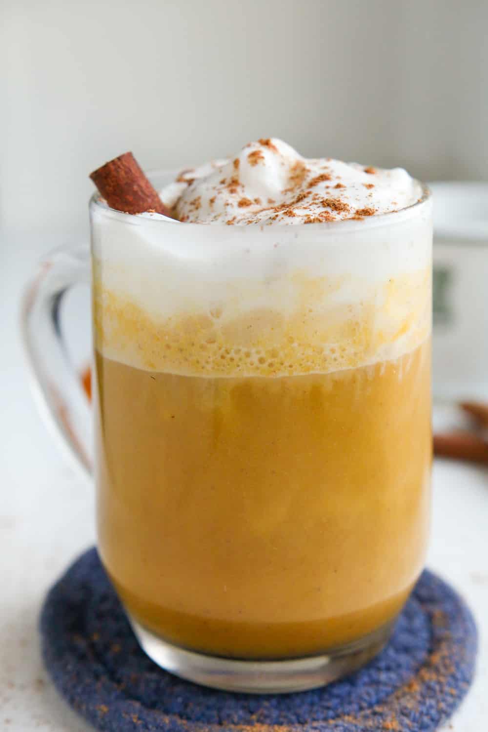 A pumpkin spice latte in a clear glass cup, and it's topped with whipped cream, a cinnamon stick, and spices.