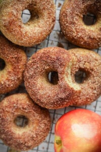 6 Donuts on a black wire rack with an apple next to them.