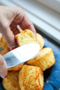 A hand holding a biscuit with a bite taken out of it and the other hand is spreading butter on it with a butter knife.