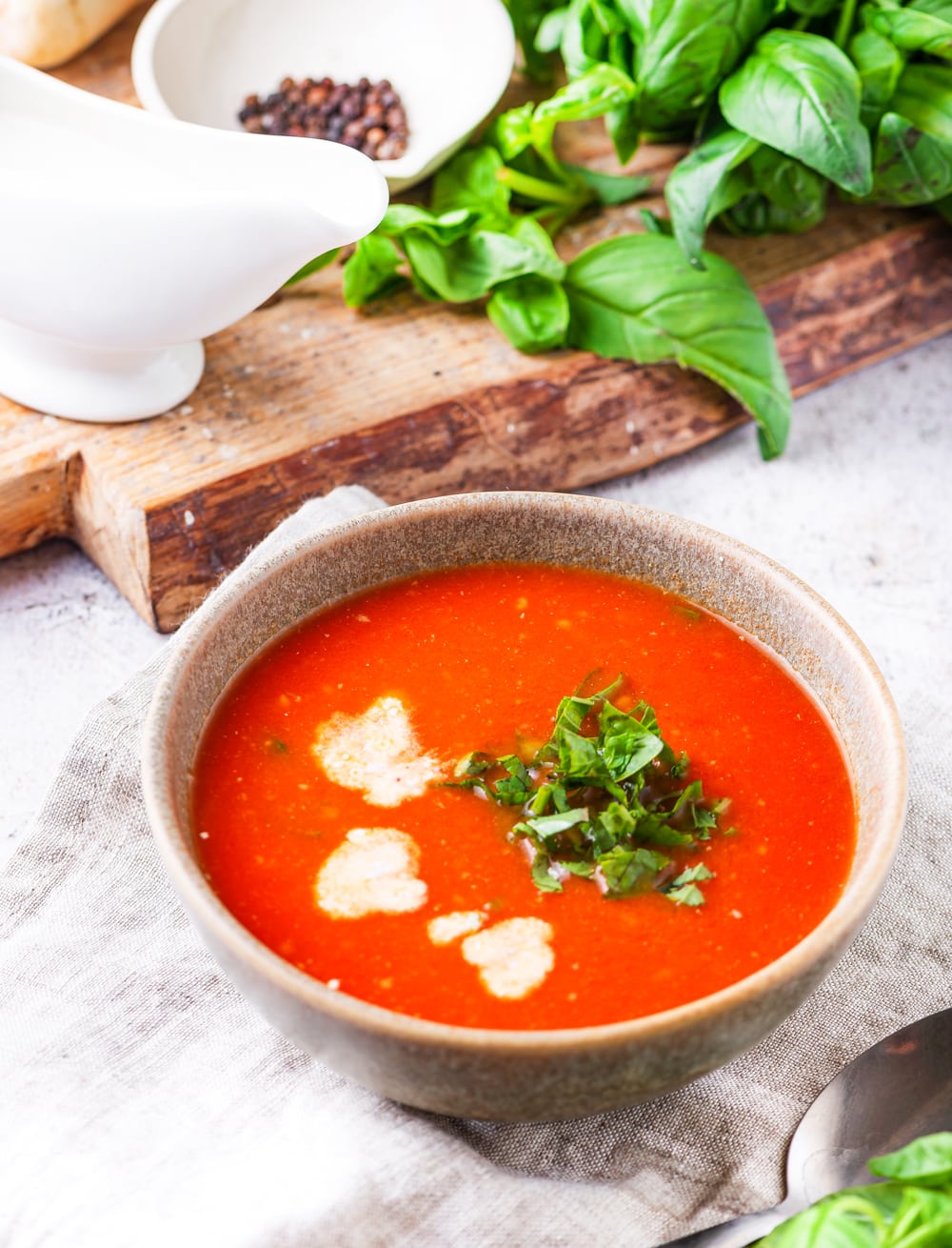 A bowl of tomato soup with a chopping board topped with basil and black peppercorns behind it.