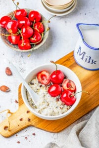 A bowl full of oatmeal, bits of dark chocolate, and cherries on a wooden cutting board.