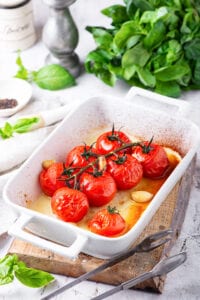 Tomatoes and garlic that have been cooked in a white baking dish with basil leaves behind it.