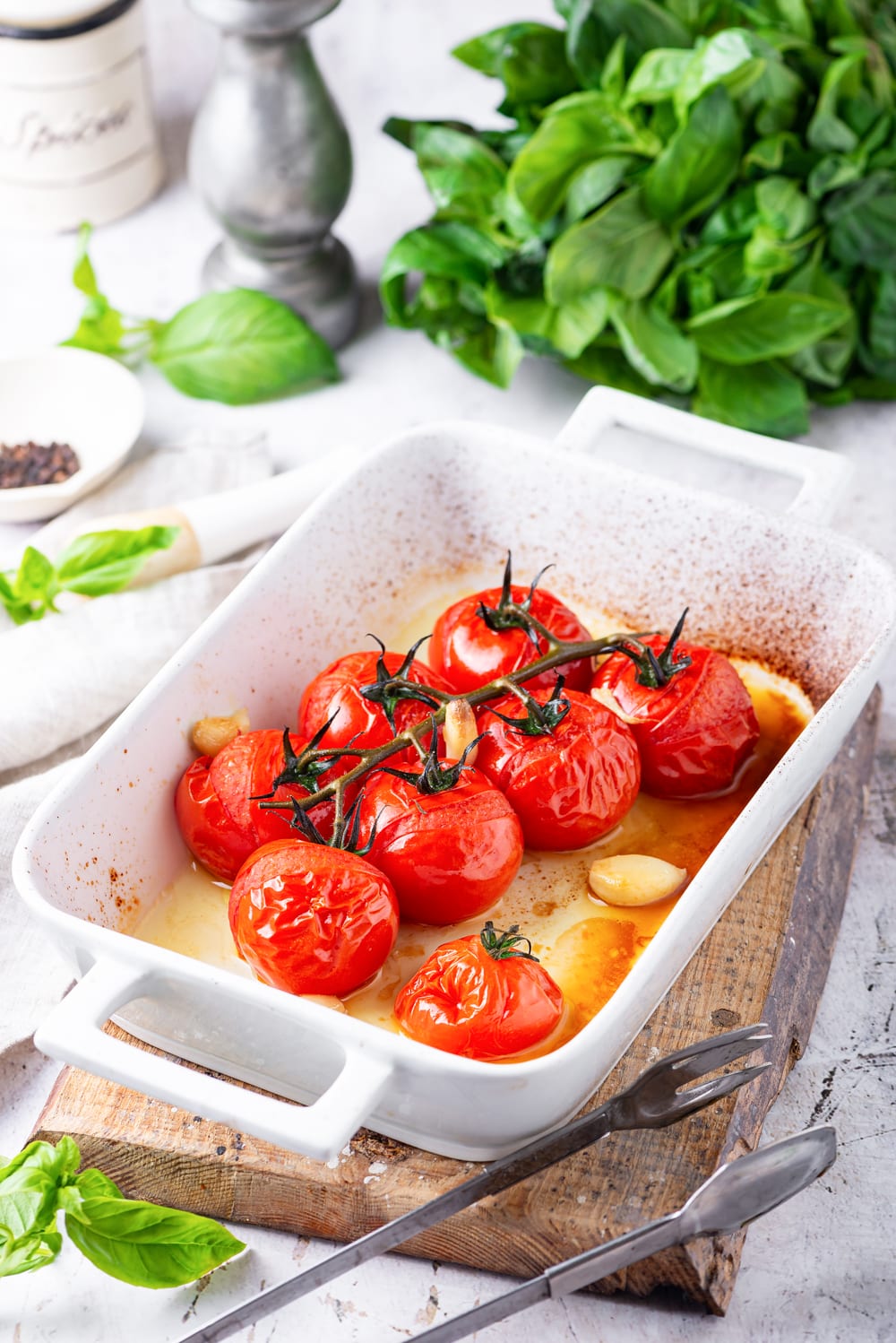 Tomatoes and garlic that have been cooked in a white baking dish with basil leaves behind it.