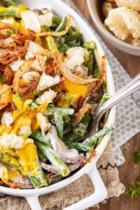 A fork placed inside half of a white casserole dish that's filled with green beans, mushrooms, onions, cheese, and pork rinds.