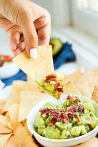 A hand holding a tortilla chip with guacamole on the tip of it.