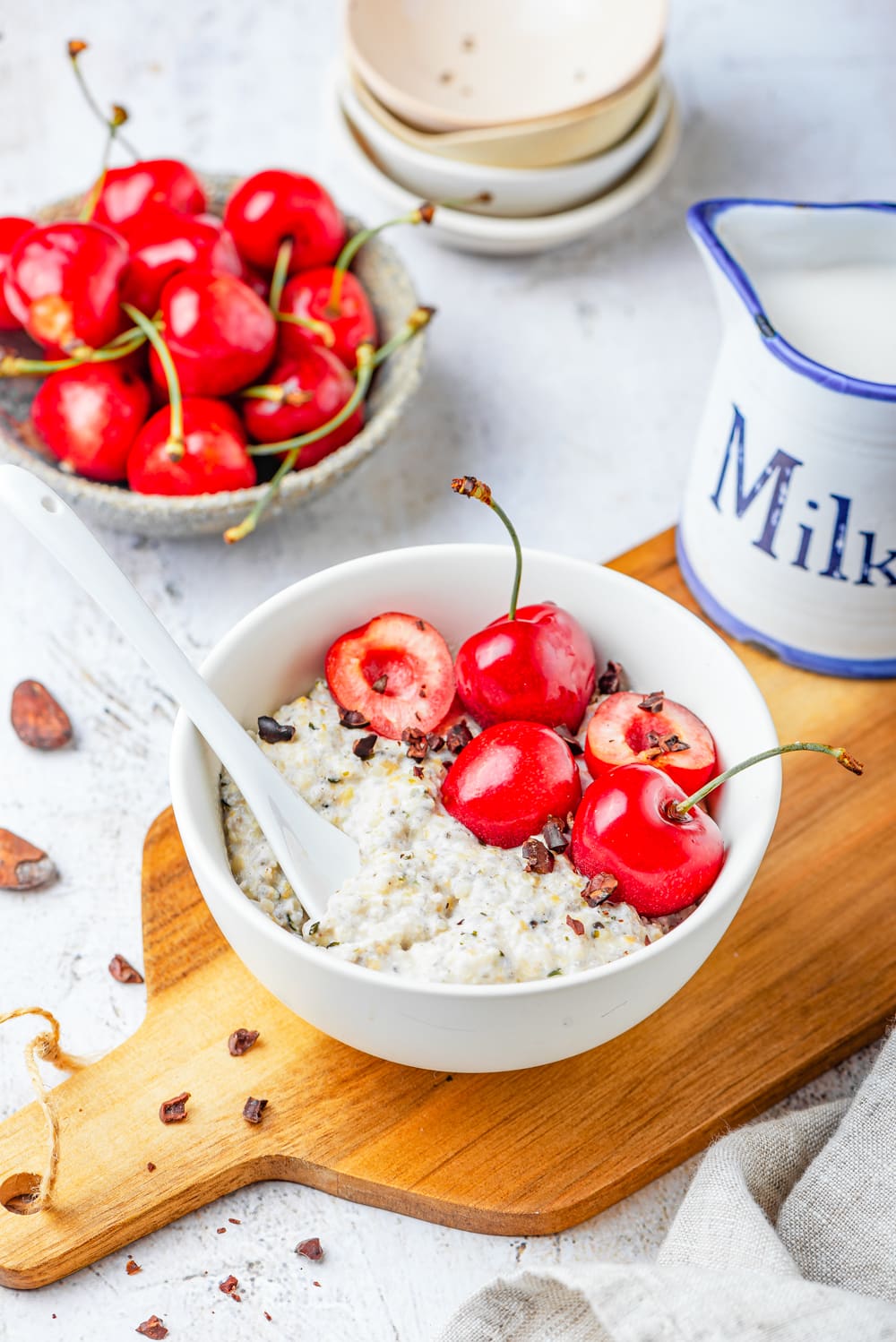 A white bowl filled with oats, bits of low carb chocolate, and cherries on a cutting board that's set on a white table.