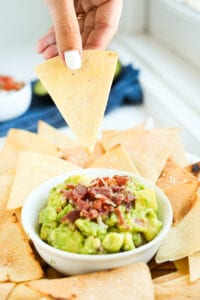 A hand holding a tortilla chip above a small of guacamole that's been topped with bacon.