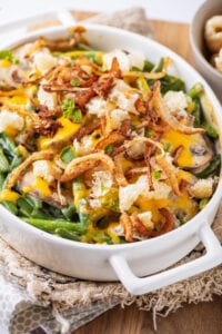 The side of a white casserole dish that's been filled with green beans, mushrooms, cheese, crispy onions, and pork rinds.