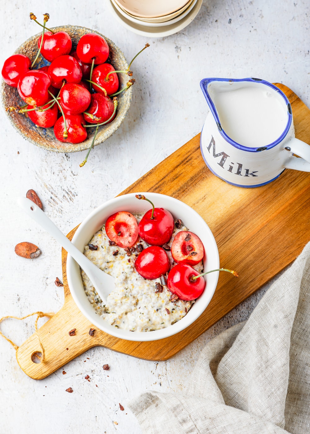 Oatmeal in a white bowl on a cutting board surrounded by a napkin, a small bowl of cherries, and a cup of milk.