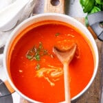 A pot of tomato soup with a spatula inside of it, and it's been set on a cutting board with basil leaves next to it.