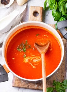 A pot of tomato soup with a spatula inside of it, and it's been set on a cutting board with basil leaves next to it.