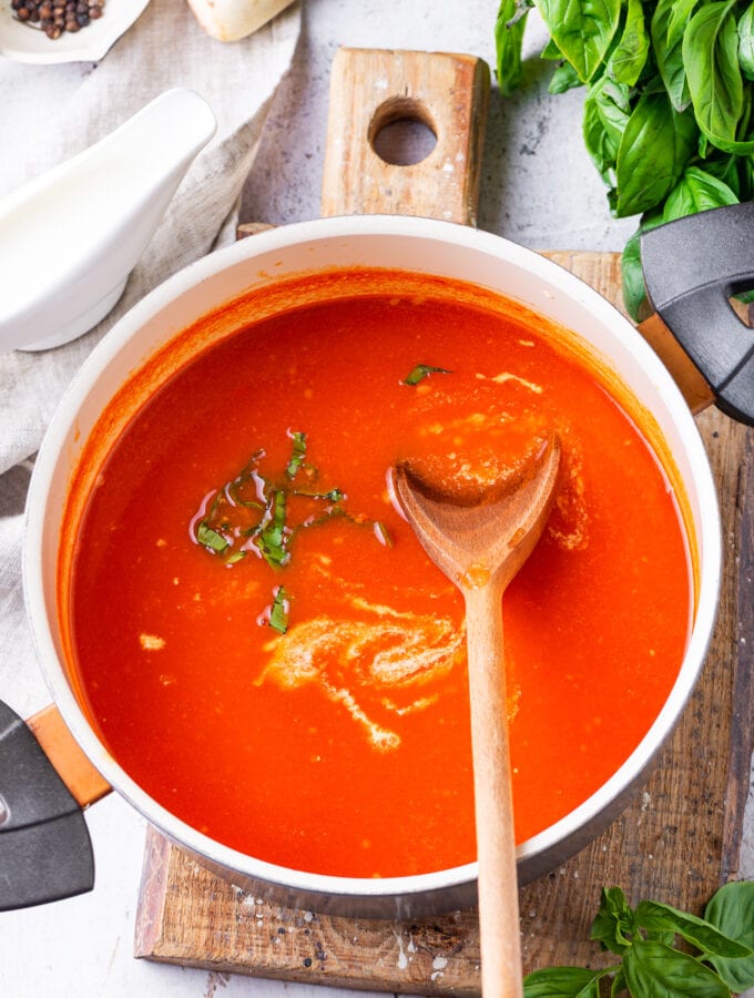 A pot of tomato soup with a spatula inside of it, and it's been set on a cutting board with basil leaves next to it.