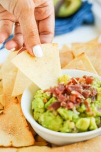 A tortilla chip being dipped into a bowl of guacamole topped with bacon.