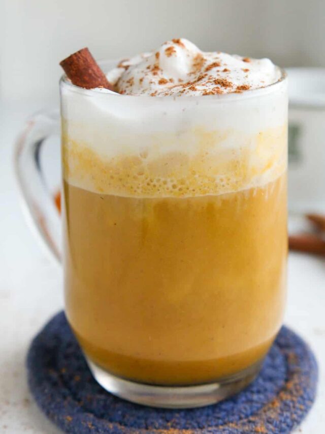 A pumpkin spice latte in a clear glass cup, and it's topped with whipped cream, a cinnamon stick, and spices.