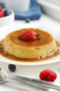 The side of a piece of flan with a strawberry on top of it and berries surrounding the plate.