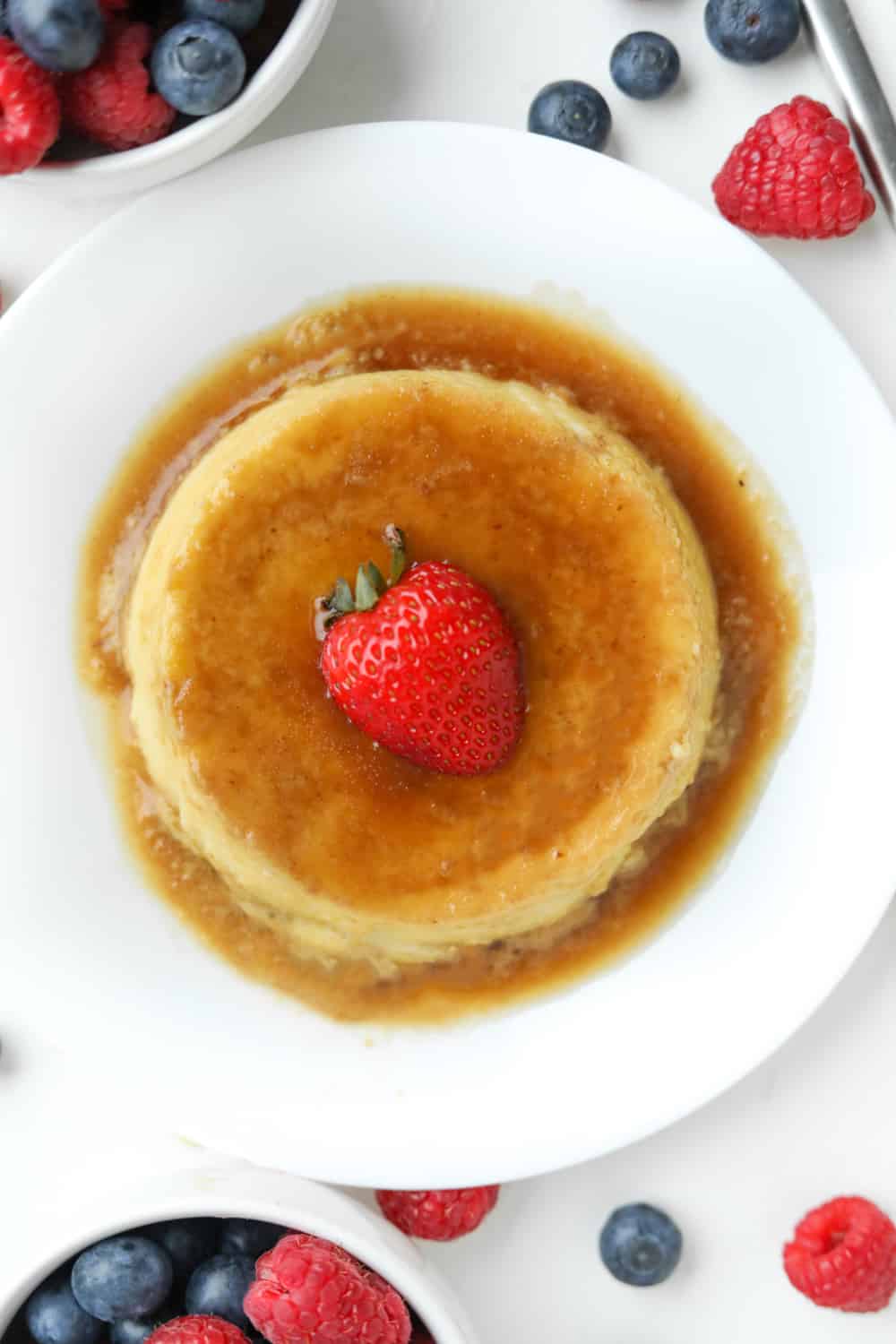 A piece of flan on a white plate set on a white table with berries all around it.