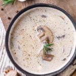 A creamy mushroom soup in a black bowl set on a wooden cutting board with sliced mushrooms next to it.