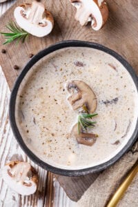 A creamy mushroom soup in a black bowl set on a wooden cutting board with sliced mushrooms next to it.