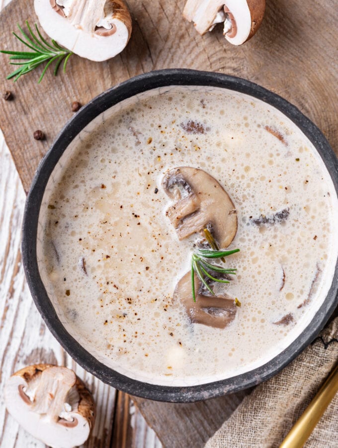 A creamy mushroom soup in a black bowl set on a wooden cutting board with sliced mushrooms next to it.