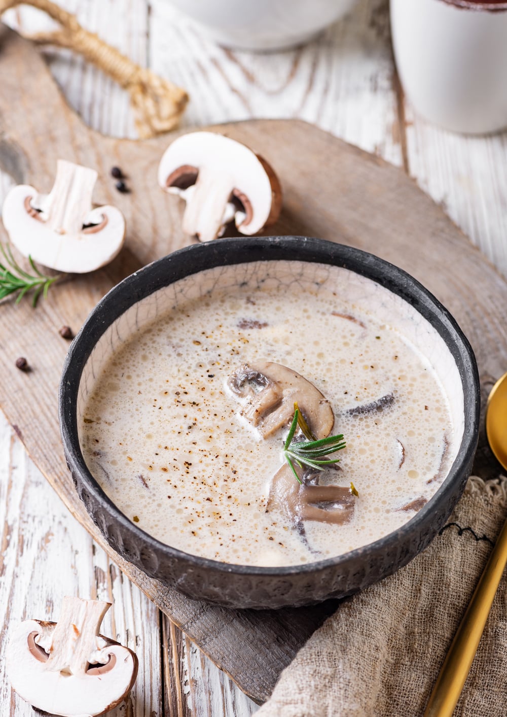 Mushroom soup garnished with a sprig of rosemary in a black bowl that's set on a wooden cutting with a golden spoon next to it.