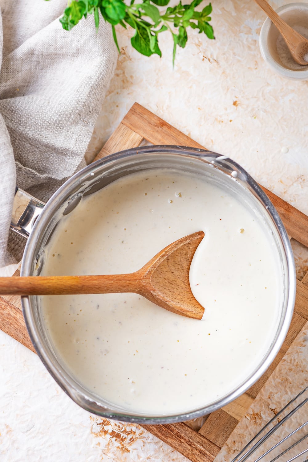 A wooden spoon placed inside of a pot filled with alfredo sauce. The pot is set on a cutting board.