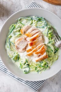 A white bowl filled with zucchini noodles, chicken, and alfredo sauce set on a blue stripped napkin.