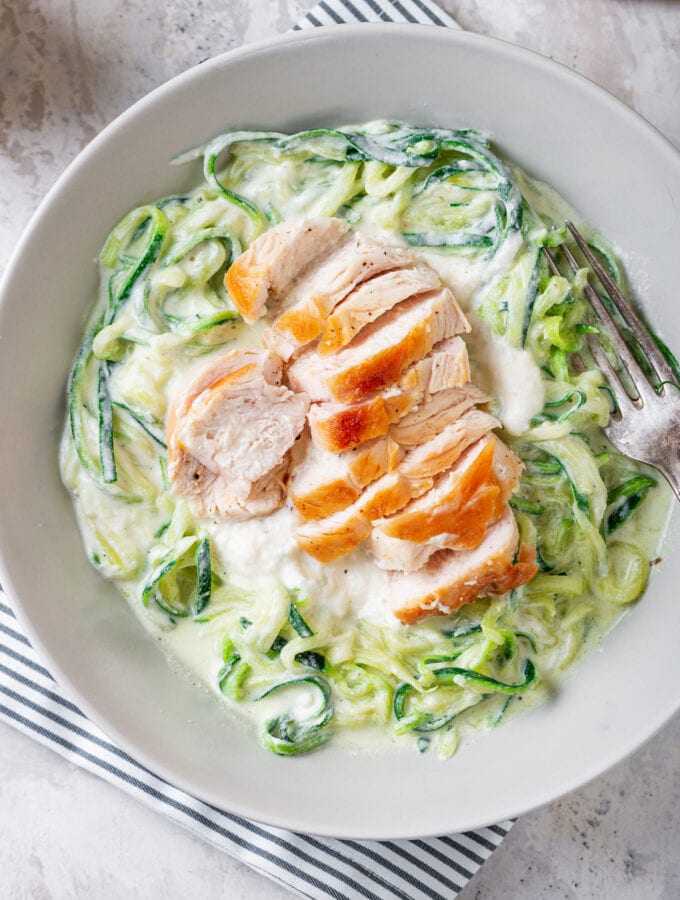 A white bowl filled with zucchini noodles, chicken, and alfredo sauce set on a blue stripped napkin.