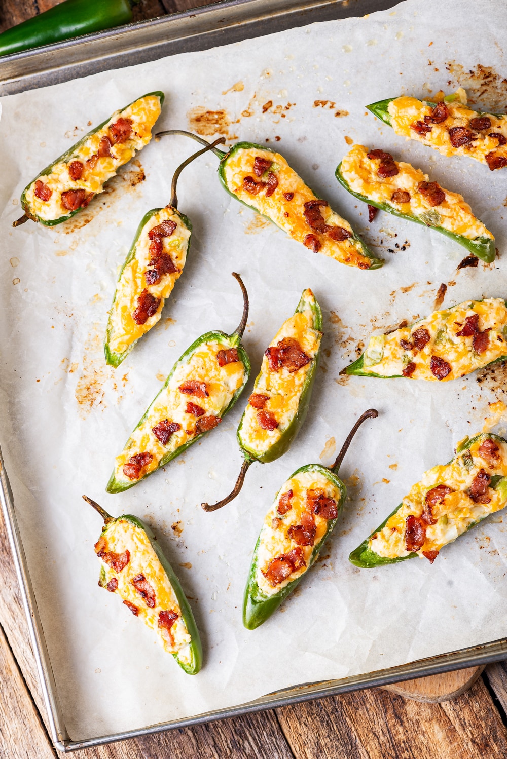 Jalapeno poppers topped with bacon on a baking sheet that's set on a wooden table.
