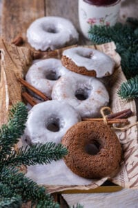 Five glazed gingerbread donuts and one plain gingerbread donut scattered on a piece of paper with cinnamon stick between the donuts. A glazed ginger bread donut is behind the piece of paper.