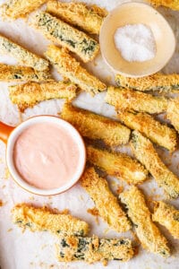 Zucchini fries on a tray lined with parchment paper and dipping sauces next to them.