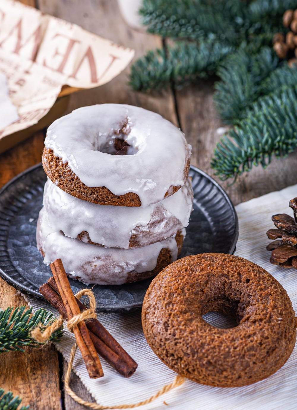 A stack of three glazed gingerbread donuts on a plate. An unglazed gingerbread donut is set to the right in front of the plate with a bundle of cinnamon to the left of the plate.