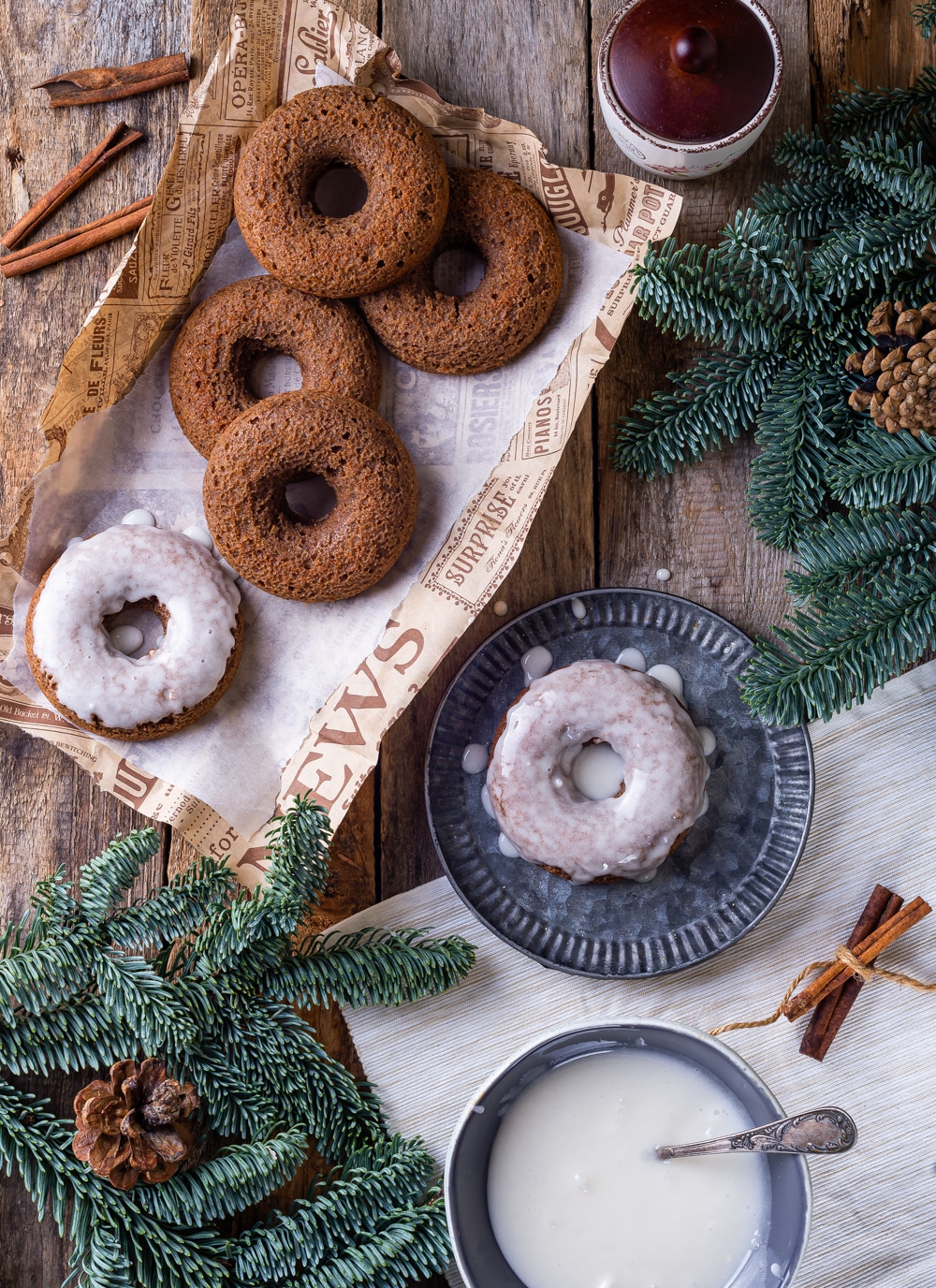 Five gingerbread donuts scattered on paper and a glazed gingerbread donut on a plate next to it. Everything is set on a wooden table with pine branches surrounding the donuts.