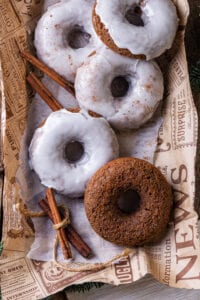 Four glazed gingerbread donuts and one plain gingerbread donut overlapping on a piece of paper. A bundle of cinnamon is next to the plain donut.