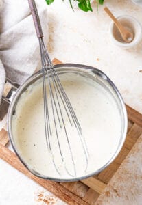 A stainless steel bowl filled with alfredo sauce. A whisk is in the sauce with the handle outside of the bowl.
