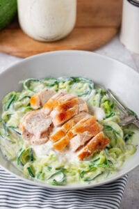 A white bowl filled with chicken on top of zucchini noodles covered in alfredo sauce.