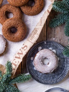 Three plain gingerbread donuts overlapping on a piece of paper. A glazed gingerbread donut is on a plate to the right of the piece of paper. Everything is set on top of a wooden table.