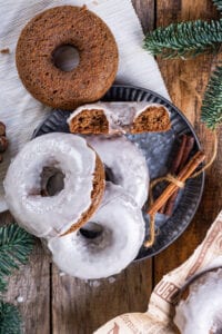 Three glazed gingerbread donuts stacked on top of one another sitting on a plate with half a gingerbread donut facing upwards behind the stack. An unglazed gingerbread donut is sitting on the edge of the plate behind the donuts.