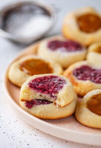 A cookie with raspberry jam filling with a bite taken out of it stacked on top of another cookie. The cookies are set on a nude plate, with more cookies behind them.