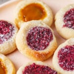 A raspberry jam filled thumbprint cookie that's set on top of a peach jam filled cookie. They are set on a plate with other cookies as well.