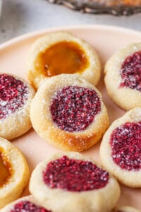 A raspberry jam filled thumbprint cookie that's set on top of a peach jam filled cookie. They are set on a plate with other cookies as well.