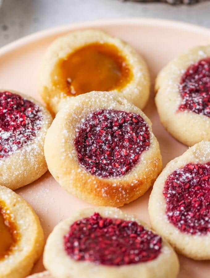 A raspberry jam filled thumbprint cookie that's set on top of a peach jam filled cookie. They are set on a plate with other cookies as well.