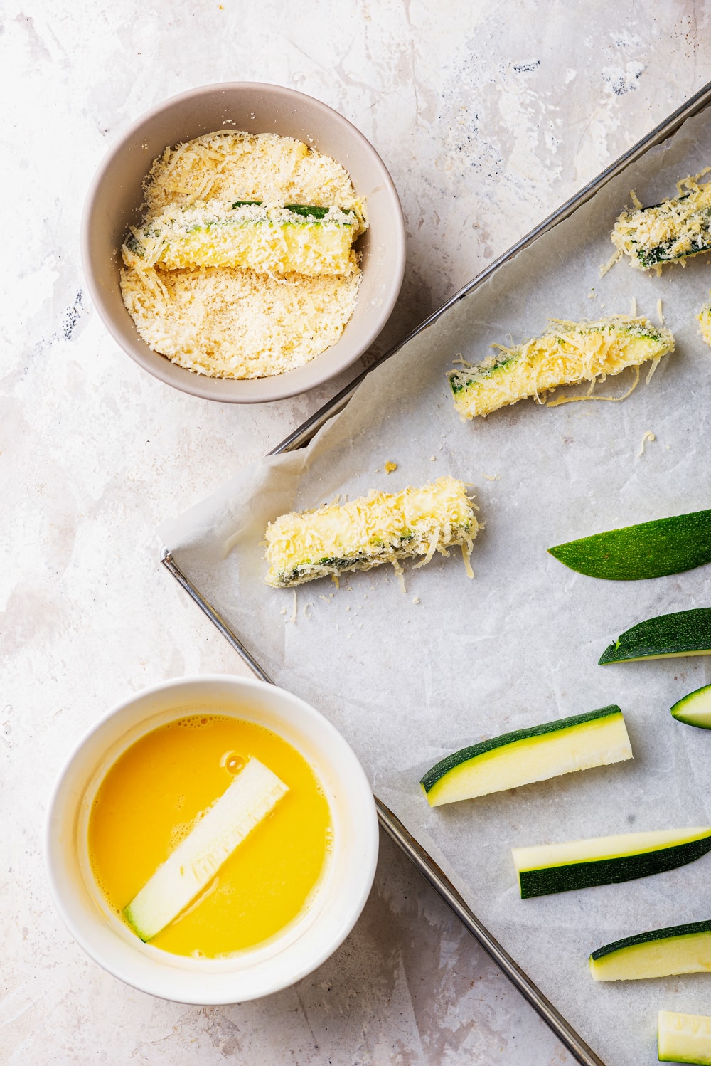 Slices of zucchini on a baking sheet. Some of the zucchini has been coated in egg and breading.