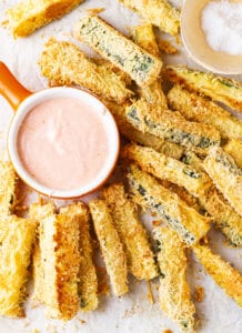 Breaded zucchini slices next to a cup of dipping sauce.