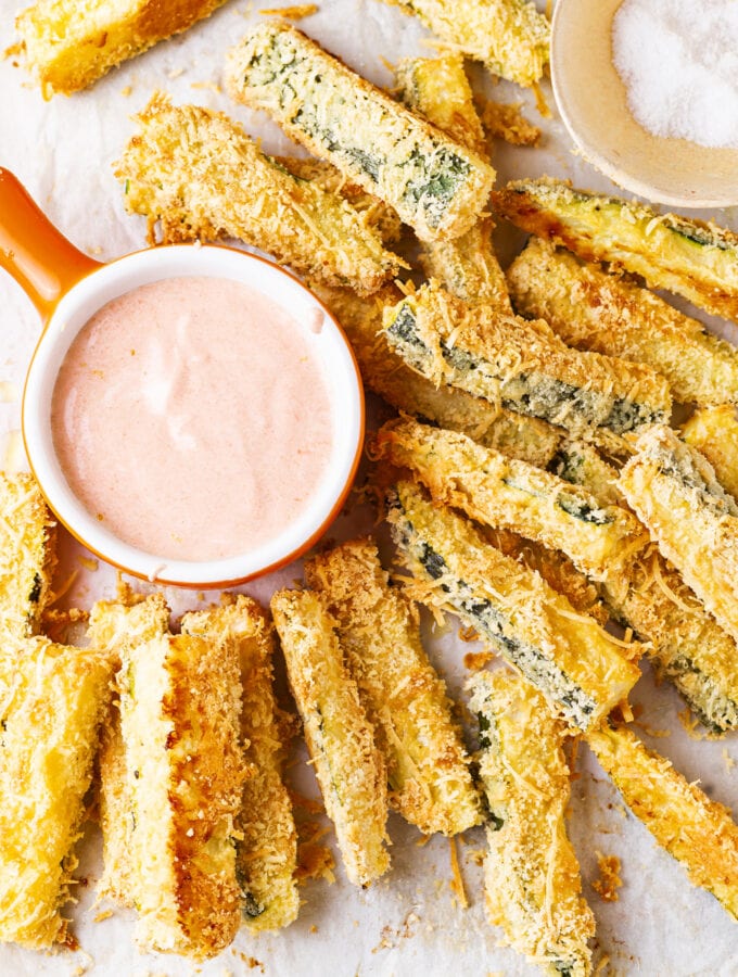 Breaded zucchini slices next to a cup of dipping sauce.