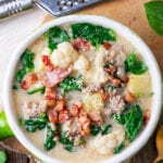 A bowl filled with soup. The soup has sausage, greens, cauliflower, and kohlrabi in it. It's also topped with bacon.