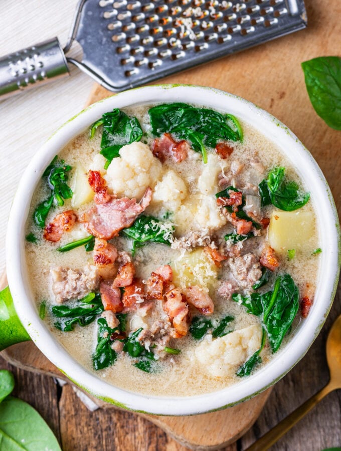 A bowl filled with soup. The soup has sausage, greens, cauliflower, and kohlrabi in it. It's also topped with bacon.