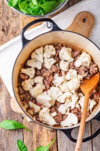 A pot with sausage, cauliflower, and a wooden spatula inside of it.